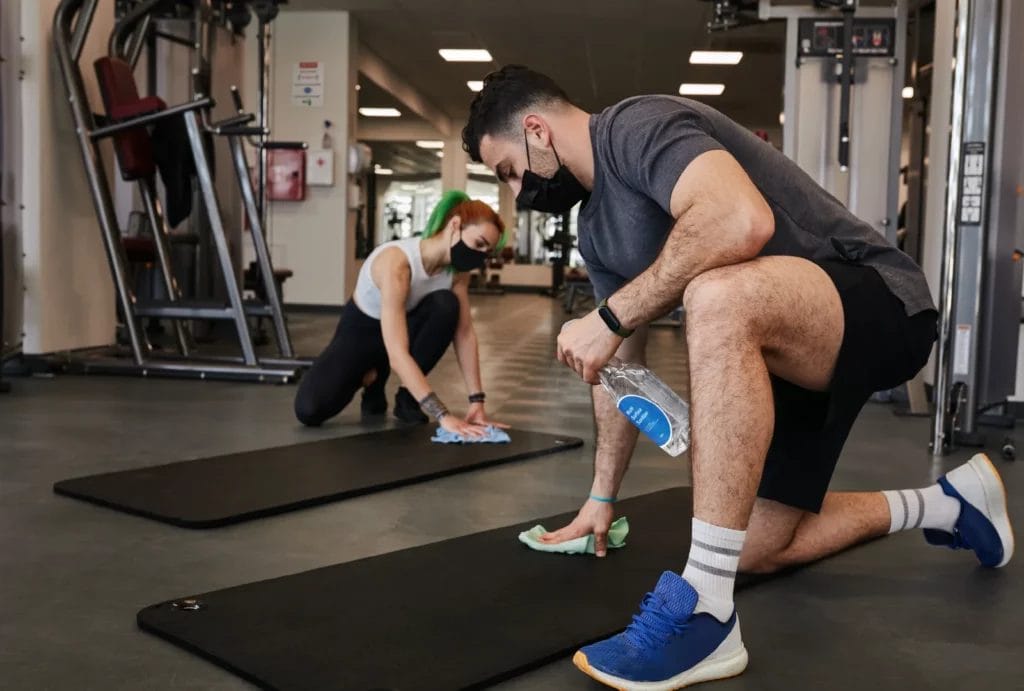 A man and a woman wearing workout clothes and face masks spray and wipe down exercise mats in the gym.