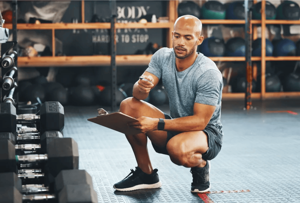 A personal trainer wearing a gray t-shirt, gray shorts, and black shoes squats in front of the weight rack and takes notes on his clipboard during an equipment assessment at the gym.