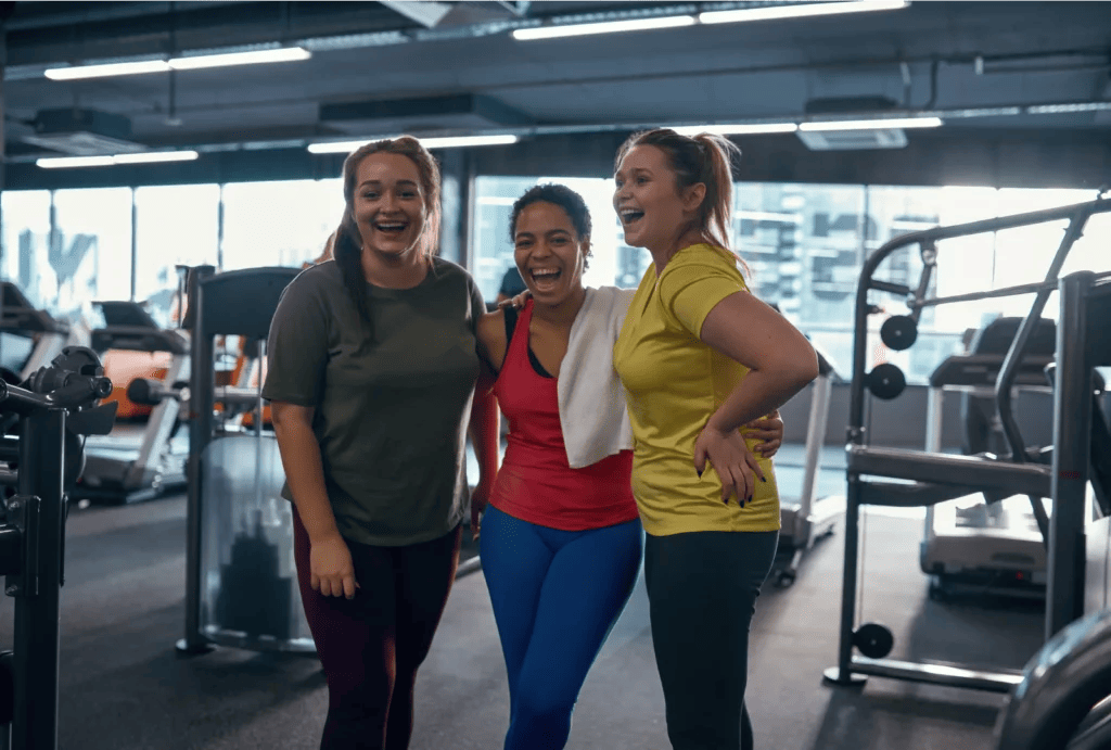 Three women, one wearing an olive T-shirt and purple leggings with a ponytail, one wearing a red tank top and blue leggings with short hair, and one wearing a yellow T-shirt and black leggings with a ponytail, laugh and pose after training at the gym.