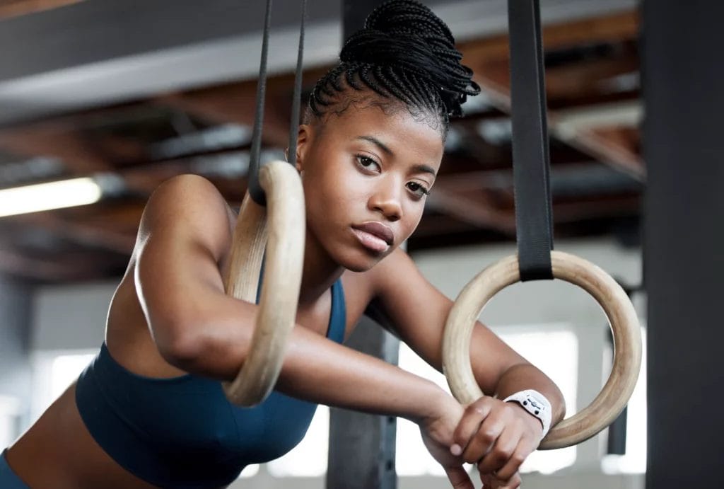 A gymnast with braided hair in a bun wearing a teal sports bra poses with her arms resting on gymnastics still rings in the gym.