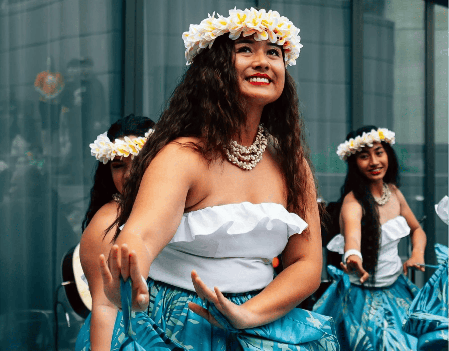 Pacific islander dancers wearing plumeria hakus, white sleeveless tops, and blue floral skirts dance in front of a glass window.