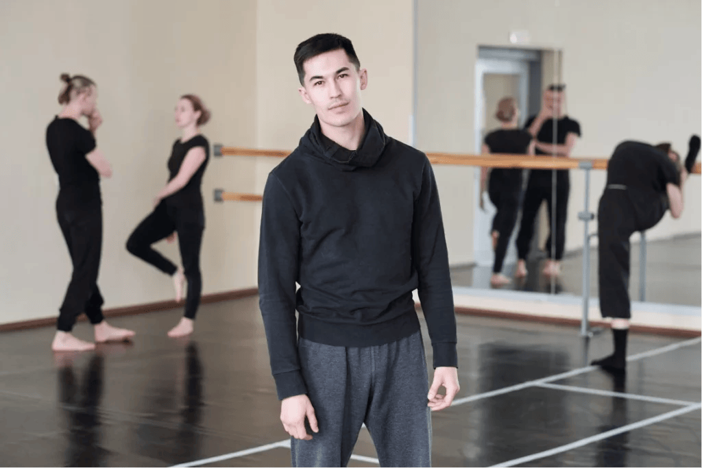 A male dance teacher wearing a black hoodie and gray sweatpants stands in front of his dance class while his students chat and stretch behind him.