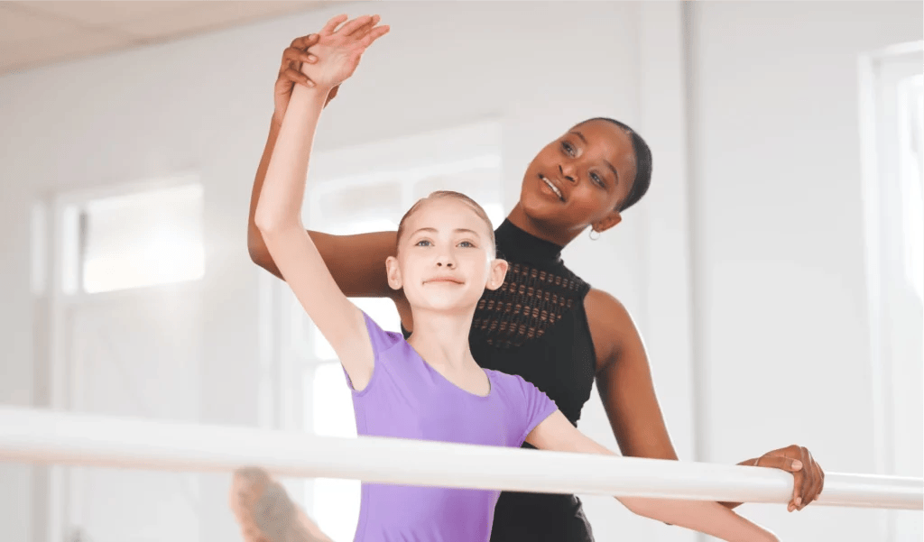 A dance teacher wearing a black leotard adjusts her student's arms at the bar during dance class.