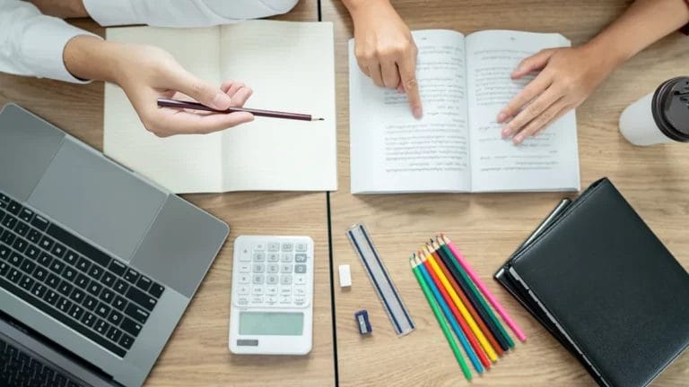 A bird's eye view of a tutor helping a student with a laptop, calculator, and notebooks spread across a desk.