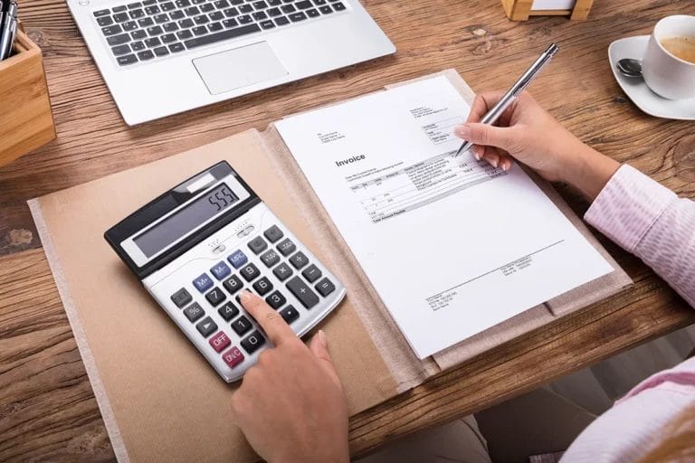 A person looking at an invoice and using a calculator on their desk.