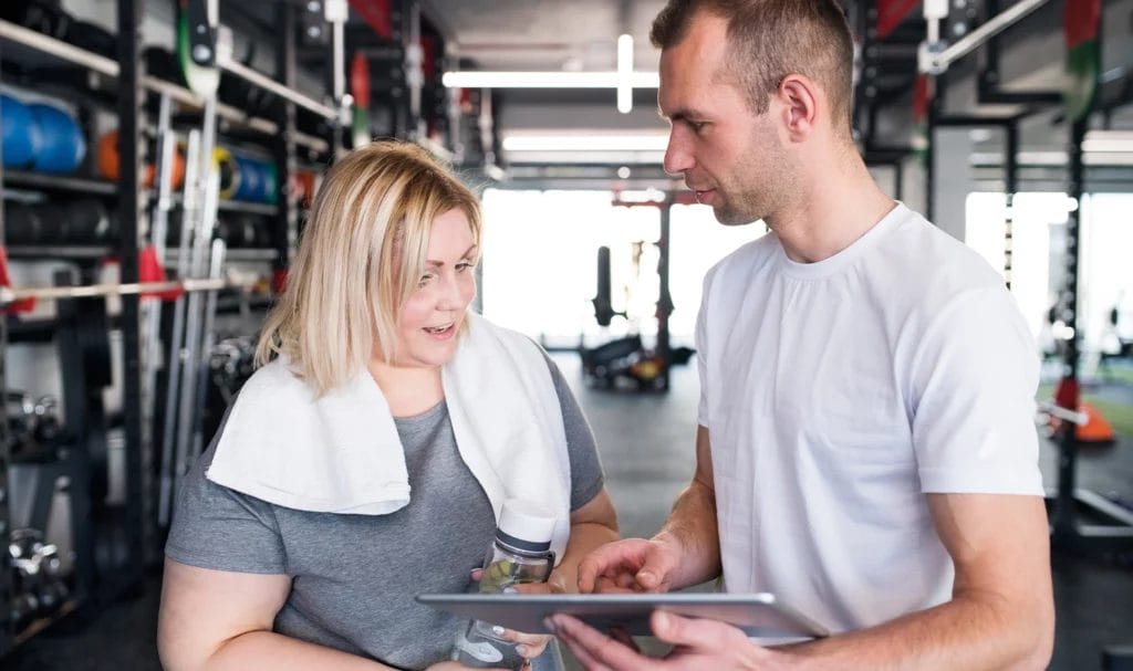 Trainer wearing a white tee, holding a tablet, reviews program with blonde client.