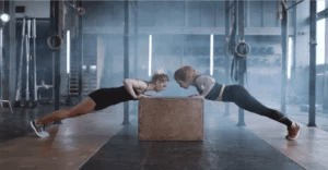 Two women wearing black activewear doing push-ups on a wooden box in a gym.