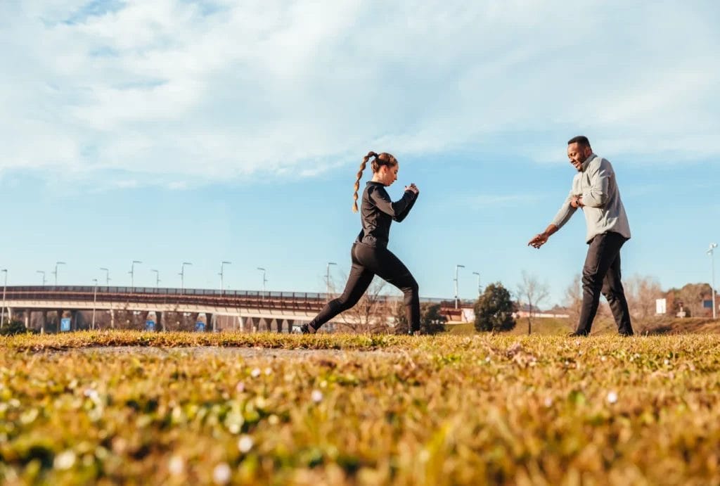 A personal trainer in a gray sweatshirt working with a client wearing black athletic clothes and a long braid, exercising in an outdoor field with a light blue sky.