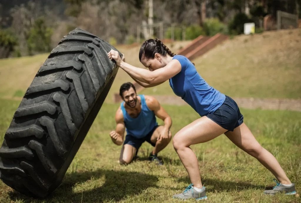 An athletic woman in a blue shirt flips a large tire while her trainer cheers her on in the background.