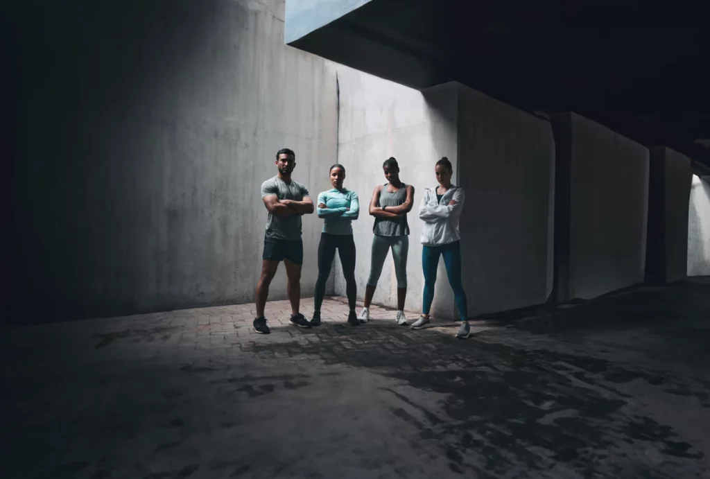 Four personal trainers wearing gray and blue athleticwear stand with their arms crossed in a shadowy industrial setting.