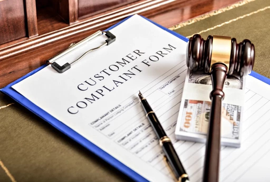 A close-up image of a "customer complaint form" on a blue clipboard with a black pen, a stack of $100 bills, and a brown gavel on a brown wooden desk with a sage-colored mat.
