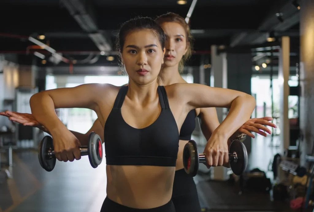 A client wearing a black sports bra holds two dumbbells while her personal trainer guides her elbows up in the gym.