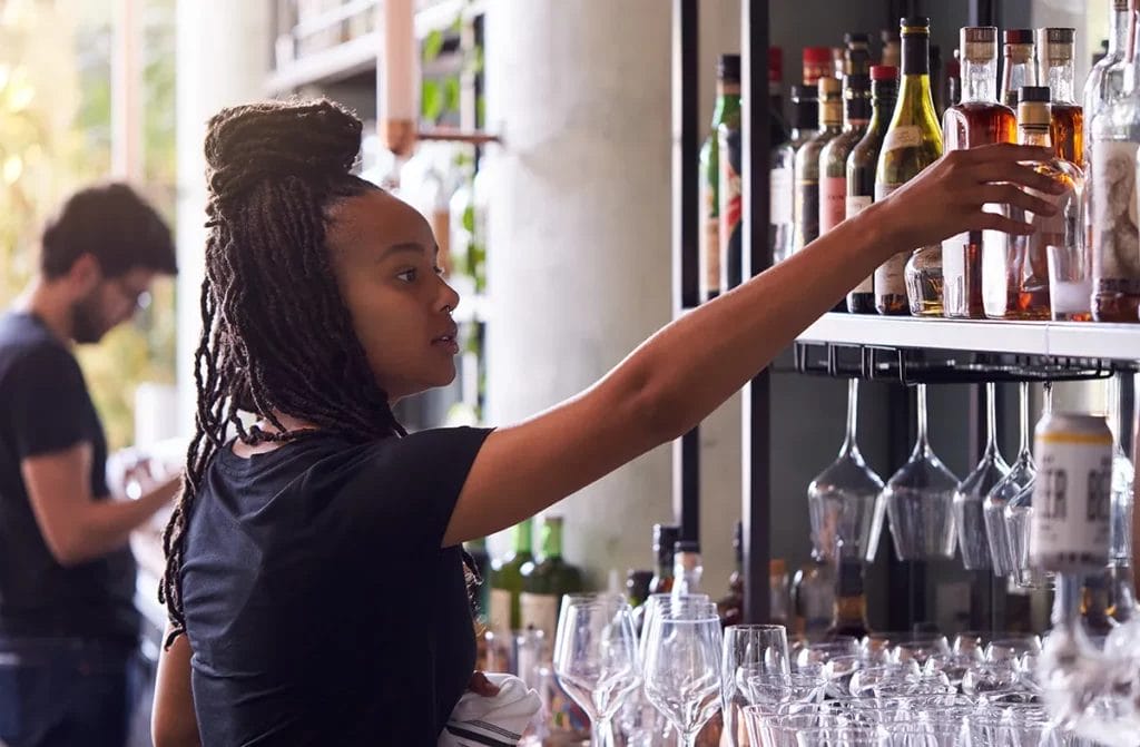 A woman reaches for a bottle of liquor while working behind a bar.