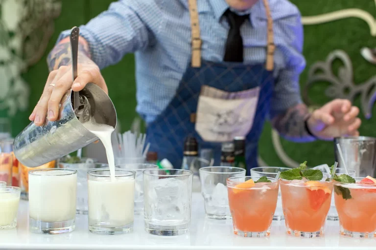 A bartender in a blue apron pours mixed drinks into glasses at an outdoor party.