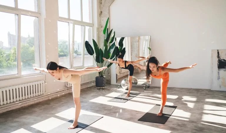 Three women practice the warrior three pose in a yoga class in a bright yoga studio with sunlight coming through the windows and a mirror, disco ball, and plant in the background.