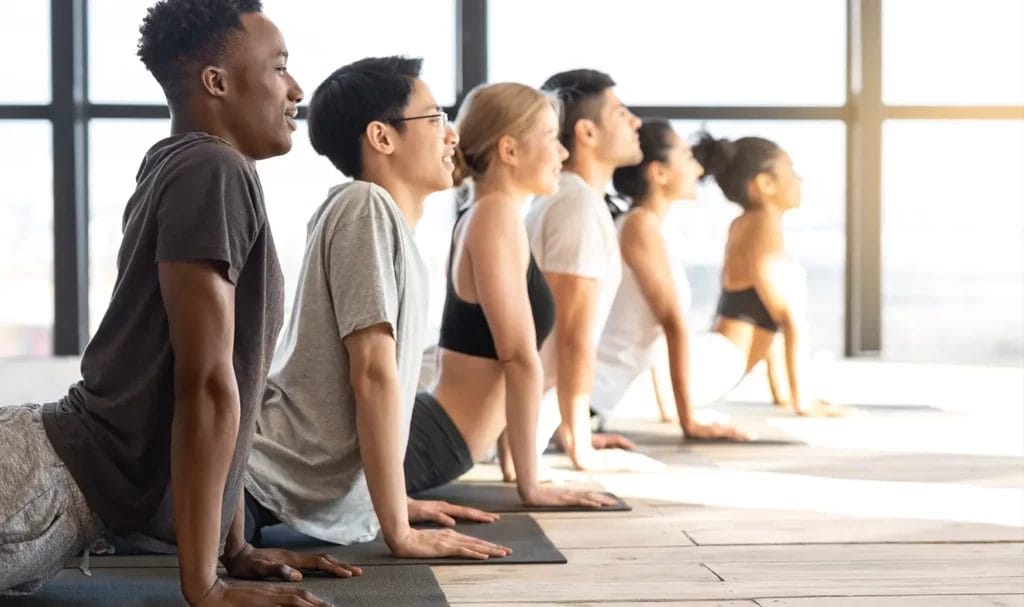 Yoga students practice cobra pose in a brightly lit yoga studio with large windows in the background.