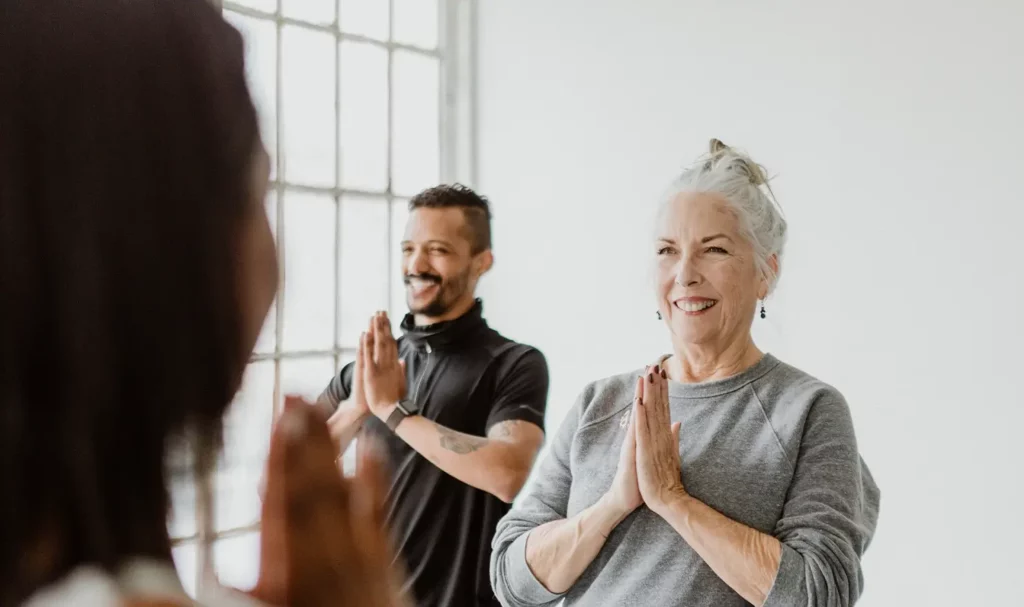 A woman and a man smile with their hands in prayer in a yoga class with the back of the teacher's head in the foreground.