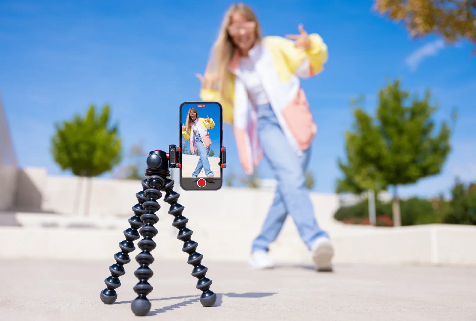 A dancer wearing a yellow jacket and jeans filming herself dancing with a smartphone balanced on a tripod.