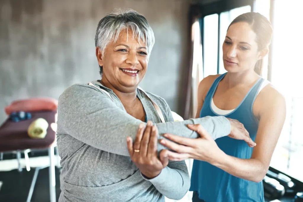 A female personal trainer wearing a blue tank top helps her female client stretch her arms in a gym.