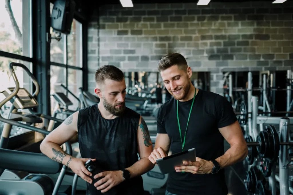 A male personal trainer wearing a black t-shirt smiles and points at his tablet as he helps a client in a gym with fitness equipment in the background.