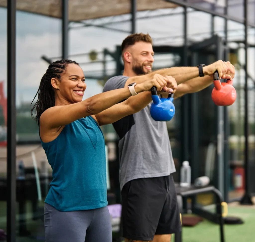 A woman wearing a teal tank top holds a blue kettlebell with both arms and a man wearing a gray t-shirt holds a red kettlebell with both arms at an outdoor fitness center.