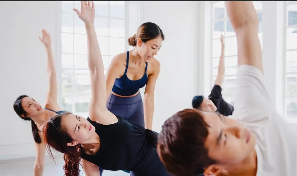 A yoga instructor stands in between students giving instructions while multiple students perform triangle pose.