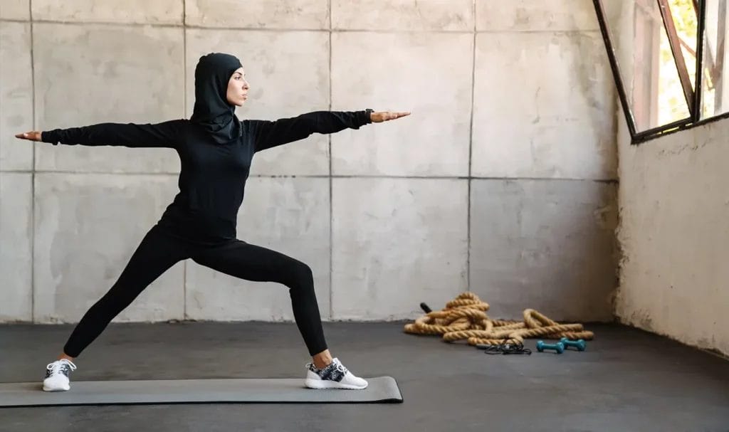 A yogi wearing black athletic clothes and a black hijab practices warrior two.
