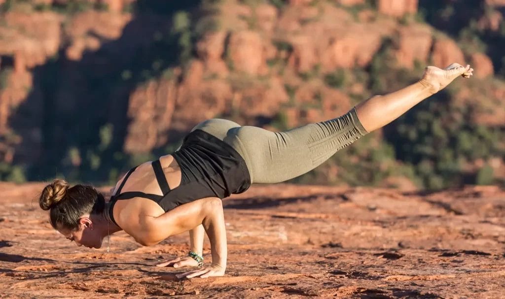 A yogi in a black tank and green pants practices a challenging yoga pose against a red rock desert background.