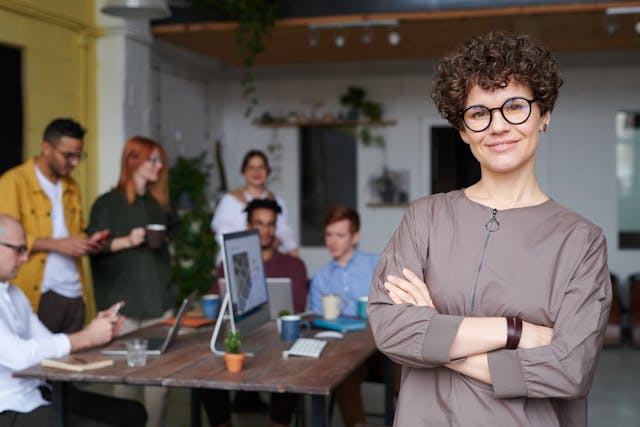 a confident woman smiles into the camera with a team behind her working on something