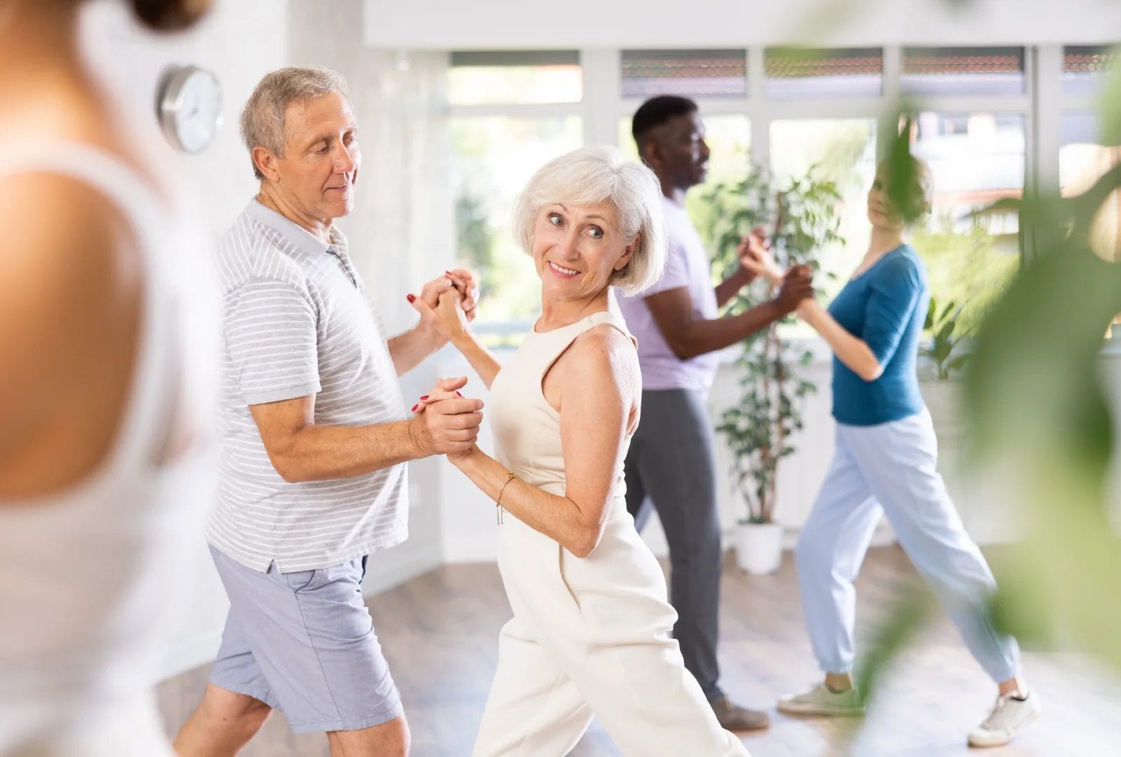 An elderly couple practices ballroom dancing in a studio with other dancers.