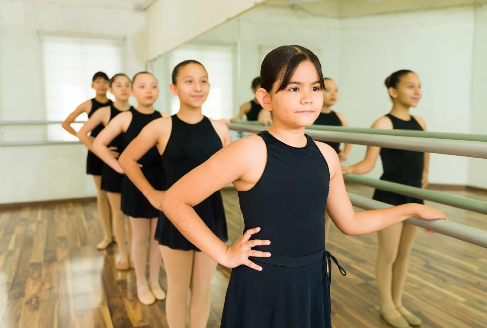 Young ballet students wearing black leotards and skirts stand at the barre and look straight forward during dance class.