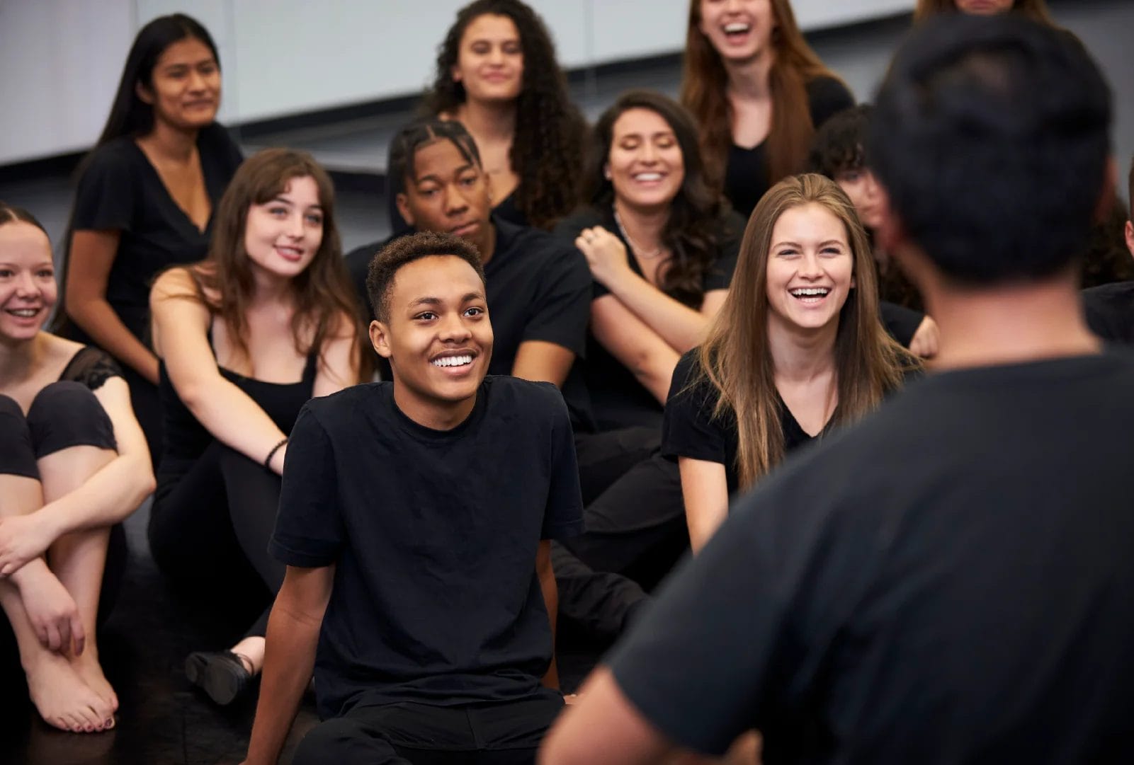 A group of dance students wearing black clothes sit on the floor, smile, and listen to their dance teacher in a dance studio.
