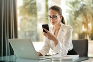 A consultant looks at notes on her phone while updating information on a laptop in an office.