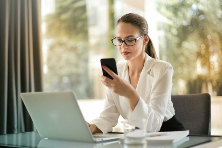 A consultant looks at notes on her phone while updating information on a laptop in an office.