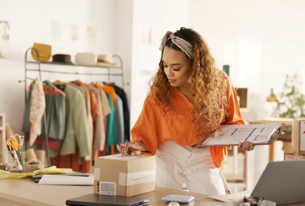 A small business designer carefully checking the label on a package with a list on a clipboard in her arms.