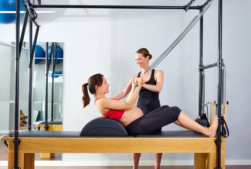 A barefoot Pilates instructor guides a pregnant client through an exercise on the Cadillac Reformer apparatus.