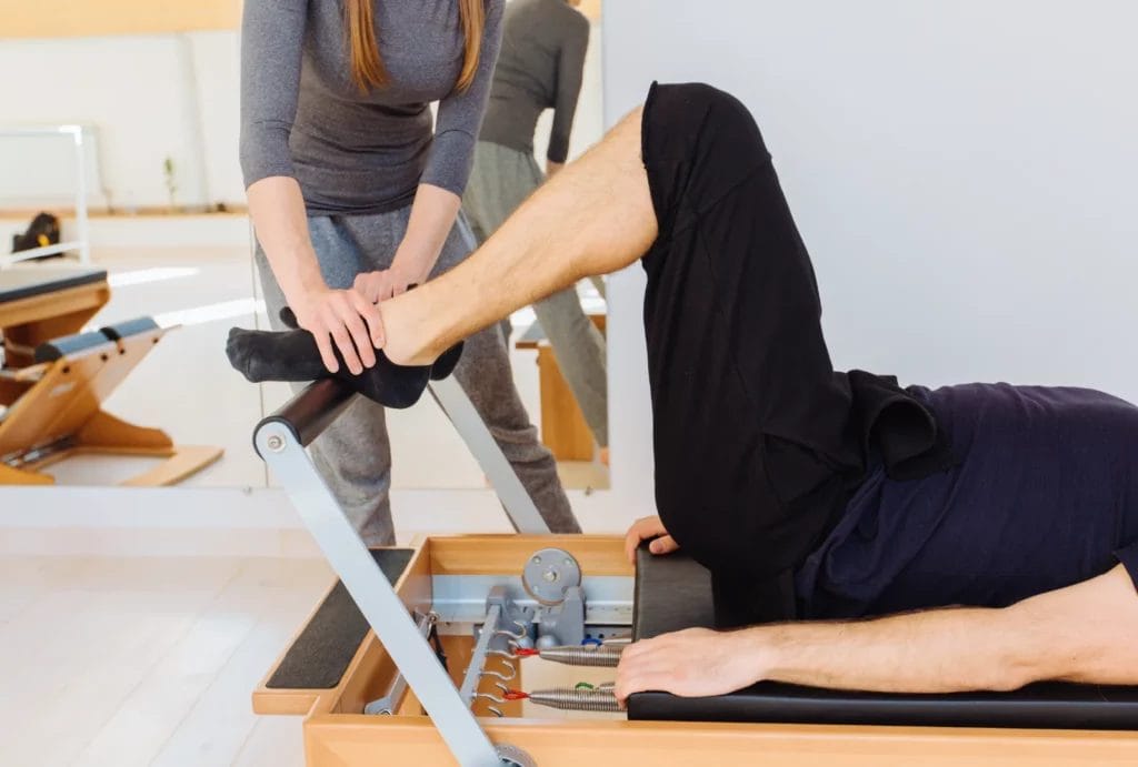 A Pilates instructor in gray athletic wear corrects a male student's foot position while on a Reformer machine.