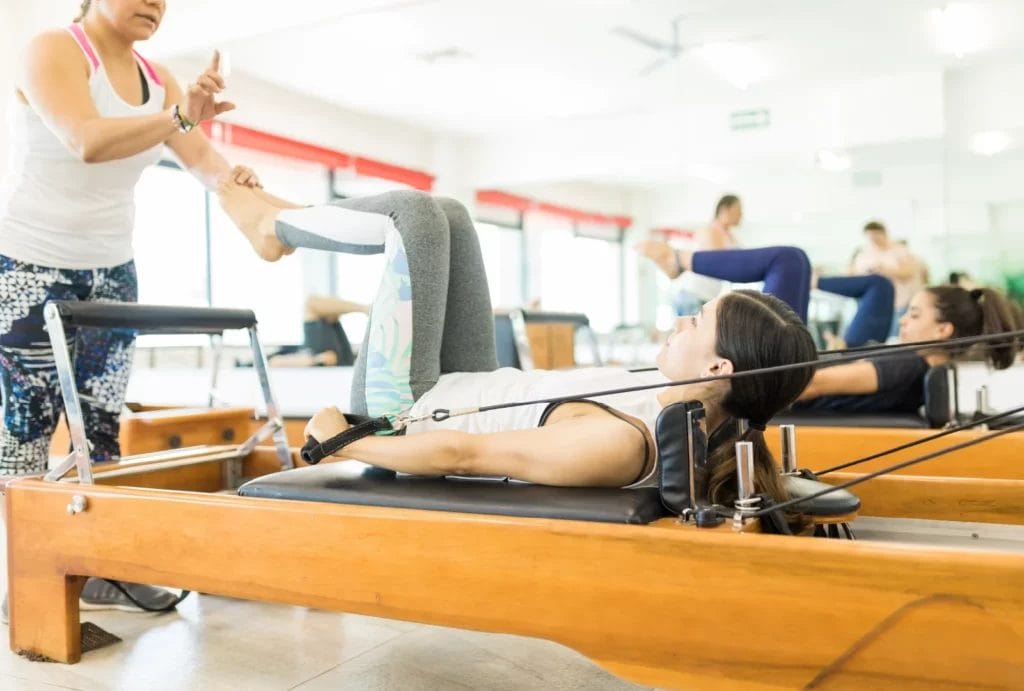 A Pilates instructor in a white tank corrects a student's foot position while on a Reformer machine.
