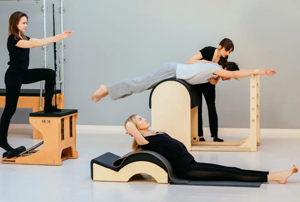 A group of instructors in training practice on different Pilates apparatus with their instructor assisting.