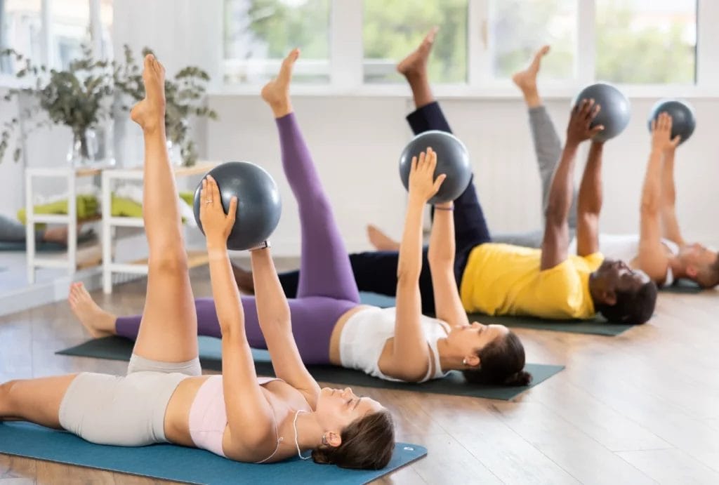 A small group of men and women participating in a Mat Pilates class with small physio balls.