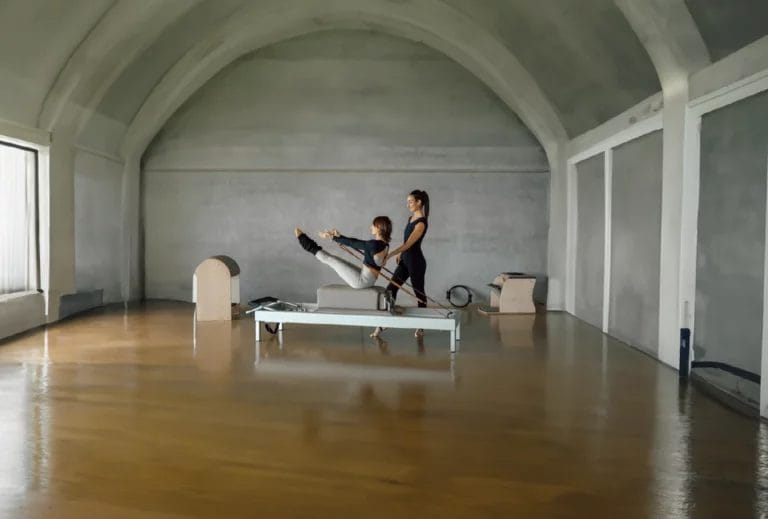A Pilates instructor in black athletic wear instructs a client through a pose on a Reformer machine in a a large open studio.
