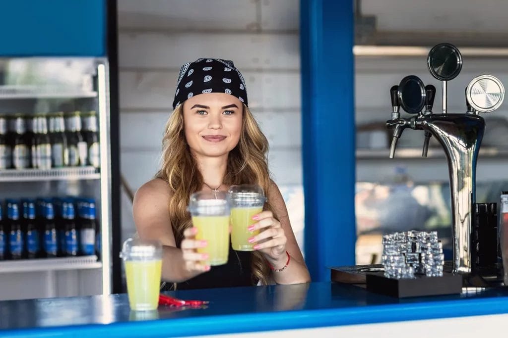 A woman wearing bandana on her head holds alcoholic beverages behind the counter at an outdoor bar.