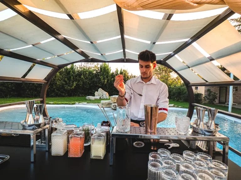 A young male bartender making a cocktail under a canopy next to an outdoor pool.