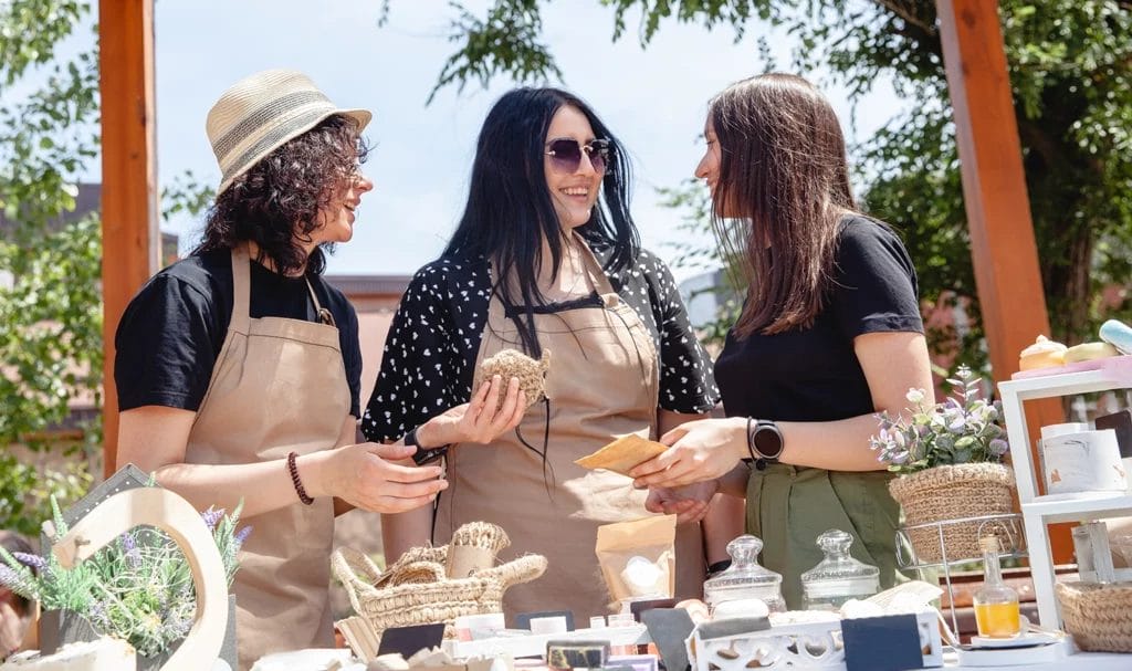 Three brunette women laughing and discussing their products they are selling at an outdoor craft fair.