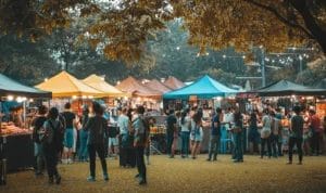 A crowd of people walking through pop-up tents at an outdoor night market at dusk.