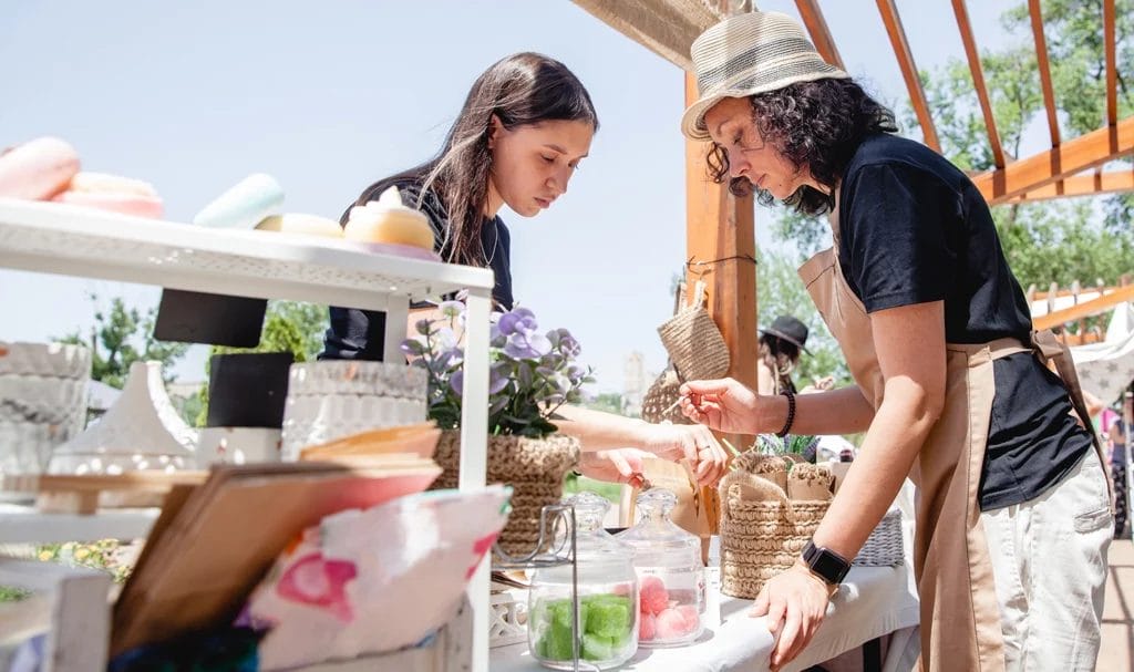 A young woman buys handmade products from a vendor, as they stand on either side of the booth at an outdoor the street fair.