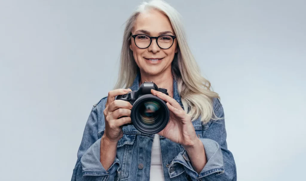 A photographer with long silver hair and black-frame eyeglasses holds up a camera and smiles while standing against a light gray backdrop.