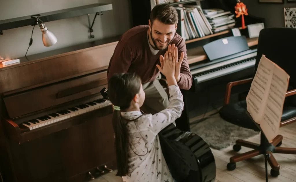 A guitar teacher high-fives a young student while they practice guitar in a music studio with a piano and keyboard in the background.