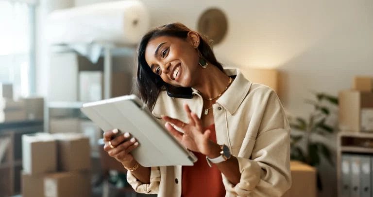 A woman wearing a beige shacket and rust-colored top smiles while talking on her smartphone, which is nestled between her ear and shoulder, and tapping on her business tablet in her home studio.
