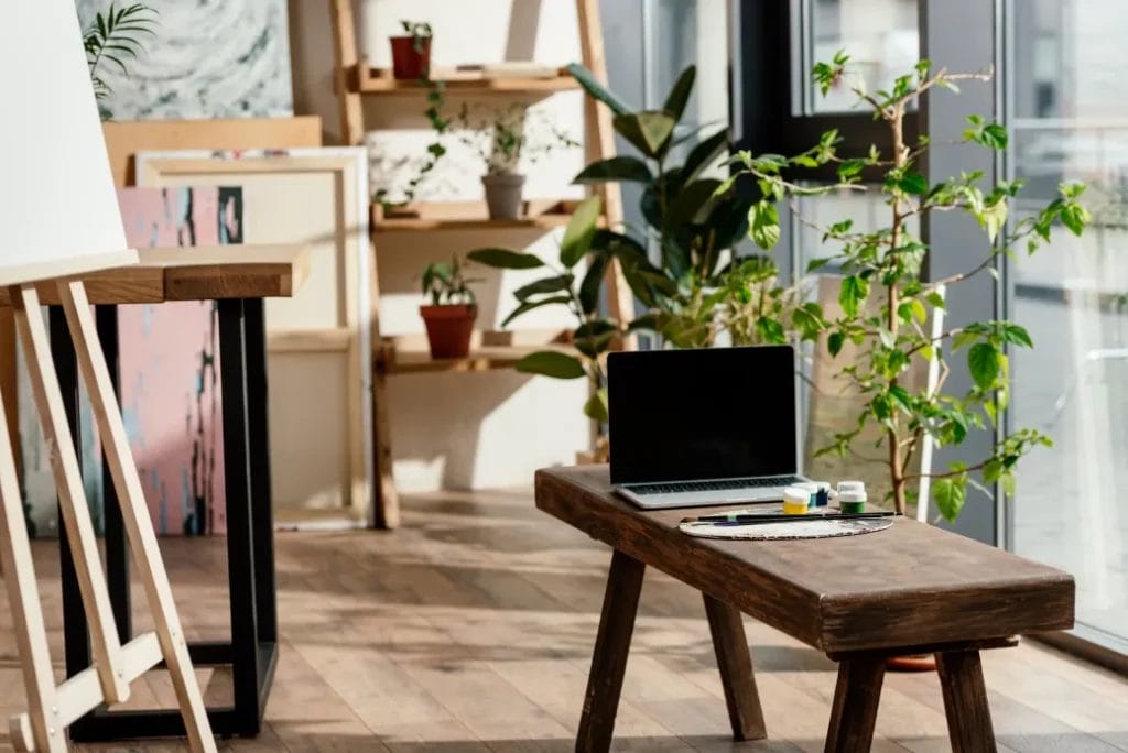 An art studio with multiple canvases, houseplants, a laptop on a wooden bench, and large glass windows.